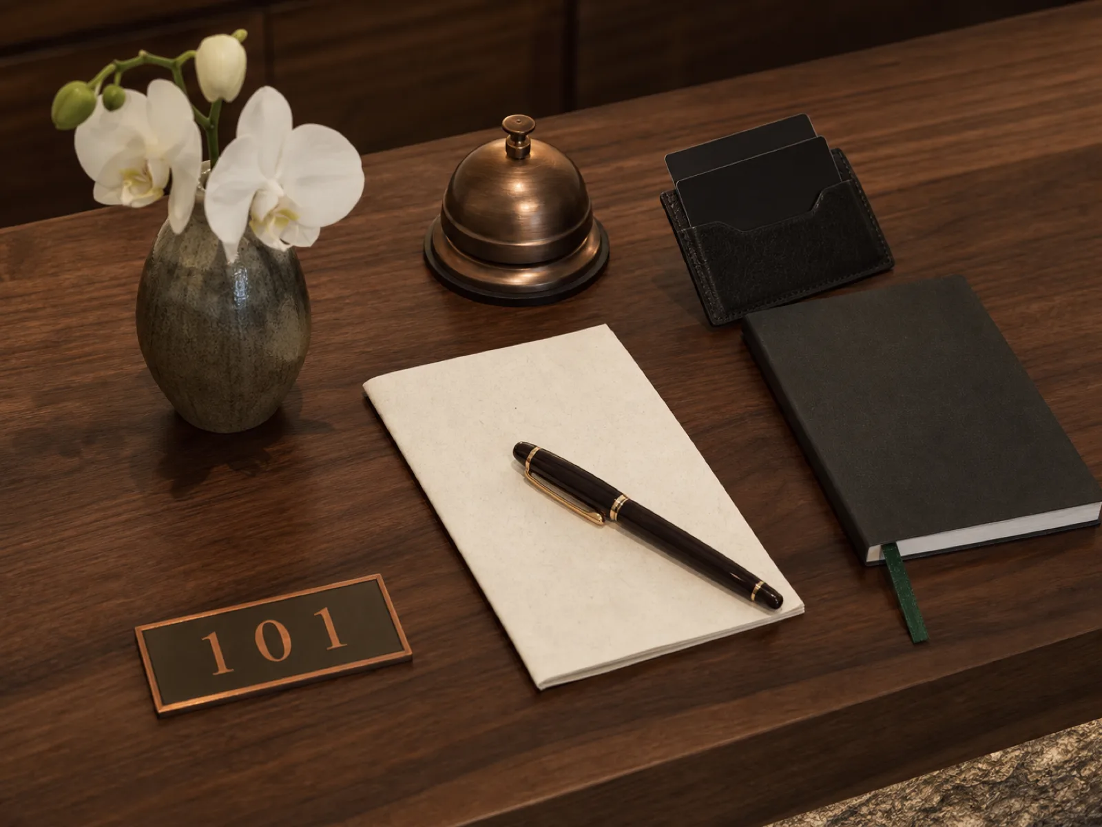 Détails de réception d'hôtel — cloche en laiton, cartes-clés, dossier client, stylo plume, orchidée fraîche — disposés sur du noyer poli